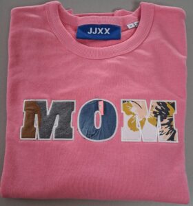 Opgevouwen roze trui waar de letters M O M geborduurd zijn met de oude babykleertjes van haar kinderen