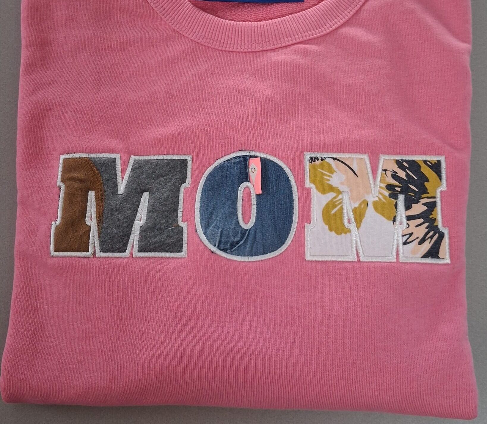 Opgevouwen roze trui waar de letters M O M geborduurd zijn met de oude babykleertjes van haar kinderen